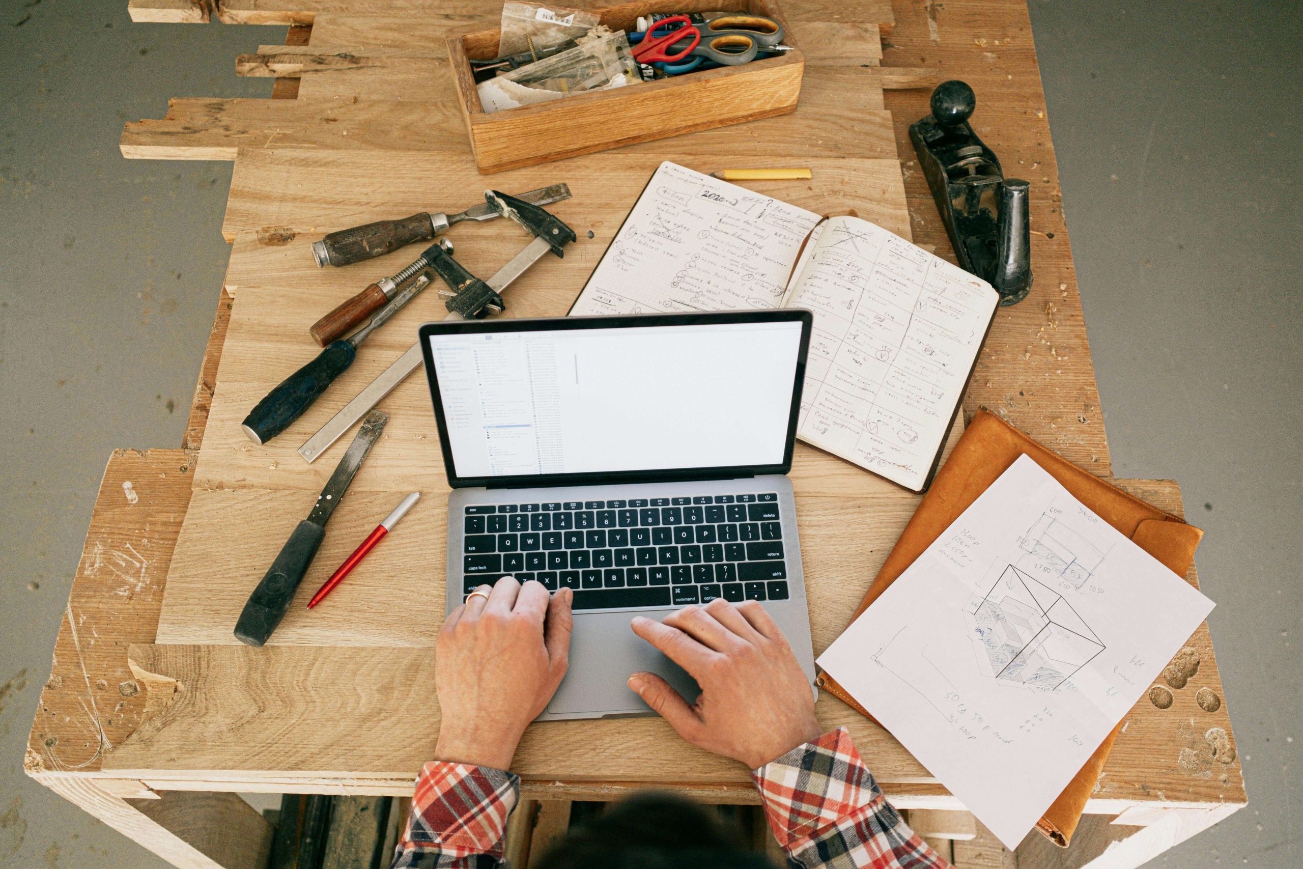 Person using a laptop on a wooden workbench with tools, notebook, and sketches.